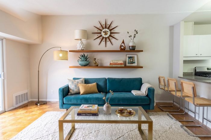Chic living room with a plush teal sofa, modern decor, and a cozy ambiance. Features stylish floating shelves, elegant lighting, and a sleek kitchen bar area.