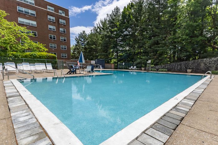 Inviting outdoor swimming pool with lounge chairs and umbrella shaded tables, set against a backdrop of lush greenery and a red brick building.