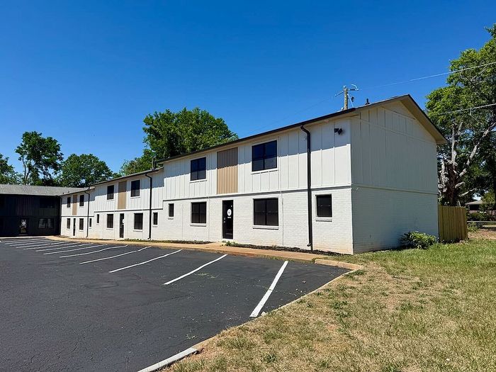 Impressive modern apartment complex with white exterior, lush greenery, and ample parking space under a clear blue sky.