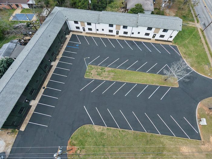 Aerial view of modern apartment complex with newly paved spacious parking lot, surrounded by lush greenery and quiet streets.