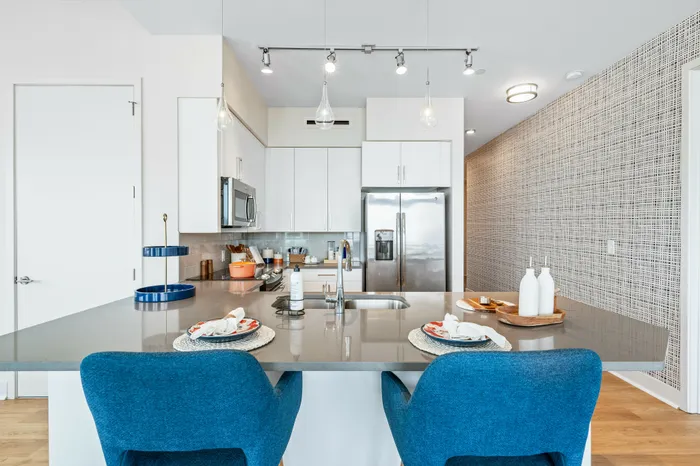 A modern kitchen in The DeSOTA with a large island for dining, featuring blue chairs, white cabinetry, and stainless steel appliances.