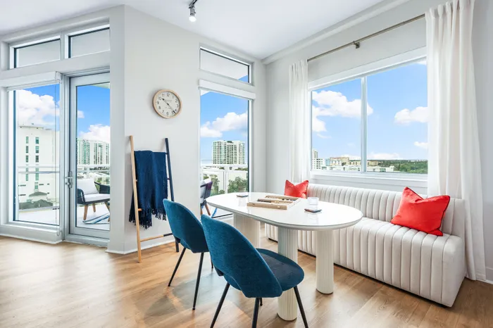 A bright dining area in The DeSOTA with a round table, modern chairs, and a bench with red pillows, with large windows overlooking the city.