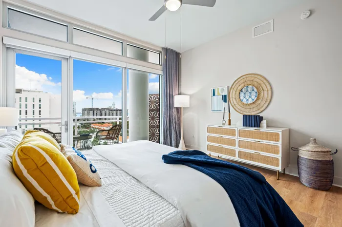 A modern bedroom in a Sarasota, FL apartment with a stylish dresser, a balcony, and large windows offering a clear view of the city.