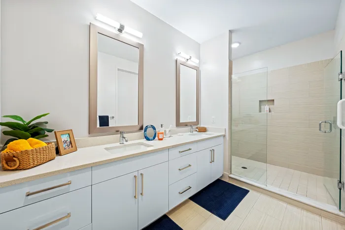 A modern bathroom at The DeSOTA with a double vanity and two mirrors, a large walk-in glass shower, and gold-colored cabinet hardware.