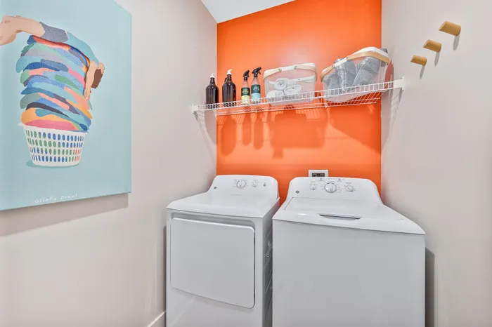 A vibrant laundry room at The DeSOTA with a white washer and dryer, a wire shelf with supplies, and a bright orange accent wall.