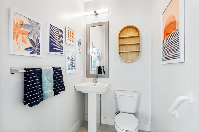 A stylish powder room at The DeSOTA with a pedestal sink, framed tropical art, a round wooden shelf, and a vibrant towel set.