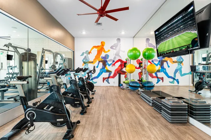 A spin and cardio room at The DeSOTA with a colorful mural of runners, spin bikes, a TV, and a large mirror on the wall.