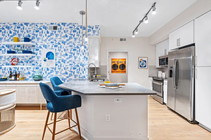 A modern kitchen in The DeSOTA with a gray island, blue bar stools, stainless steel appliances, and a bold blue patterned wall.