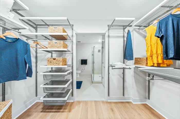 A spacious walk-in closet in a The DeSOTA apartment with a modern shelving system, a light wood floor, and an open view into a bathroom.