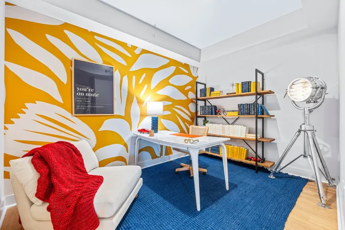 A unique home office in a The DeSOTA apartment with a bold yellow patterned wall, a desk, a bookshelf, and a large vintage-style light.