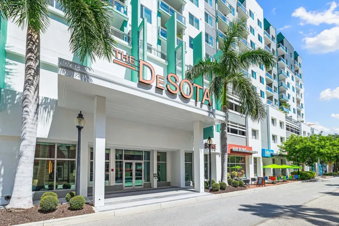 The main entrance of The DeSOTA apartment building, with its name prominently displayed above the glass doors, framed by palm trees and street-level shops.