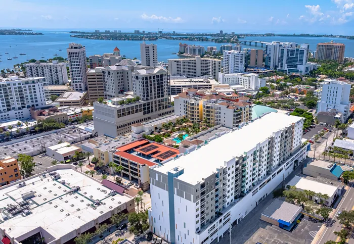 An aerial view of the vibrant downtown Sarasota skyline, showcasing numerous high-rise buildings, with a scenic bay and bridge in the background.