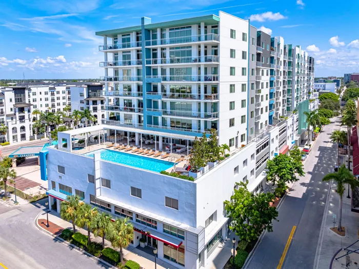 An aerial view of a modern multi-story apartment building in Sarasota, FL, with a rooftop pool and a sleek white and blue facade.