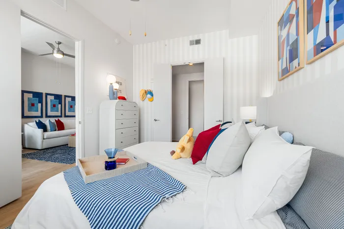 A modern bedroom in a The DeSOTA apartment with striped walls, a bed with colorful pillows, and a view into the living room.