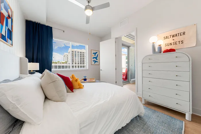 A modern bedroom in a The DeSOTA apartment with a bed, dresser, and a window with a scenic view. An open door leads to a balcony.