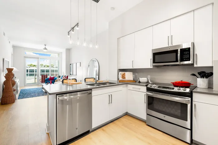 An open-concept living and kitchen area in a The DeSOTA apartment with modern white cabinets, stainless steel appliances, and a balcony.
