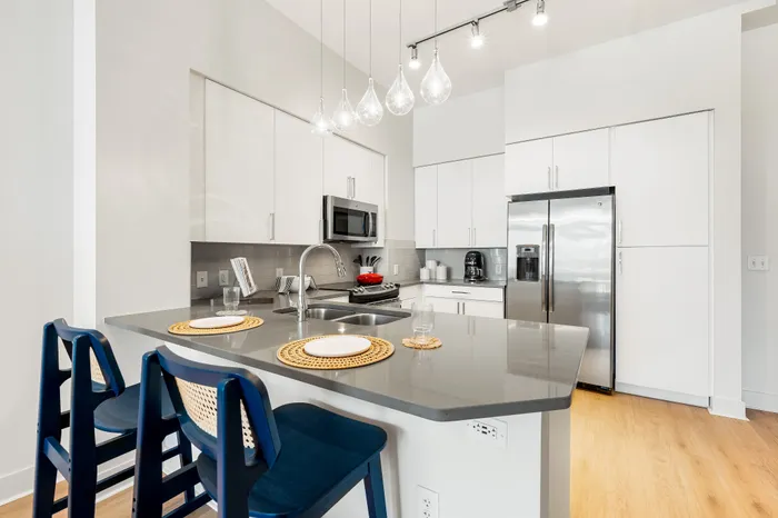 A modern kitchen at The DeSOTA with a gray island, blue bar stools, sleek white cabinets, and stainless steel appliances.