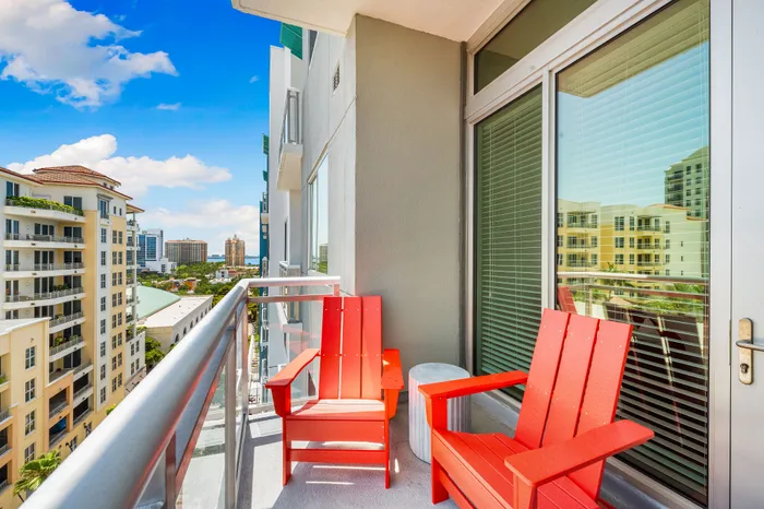 A furnished balcony in a Sarasota, FL apartment with two bright red chairs and a glass railing, offering a view of the city.