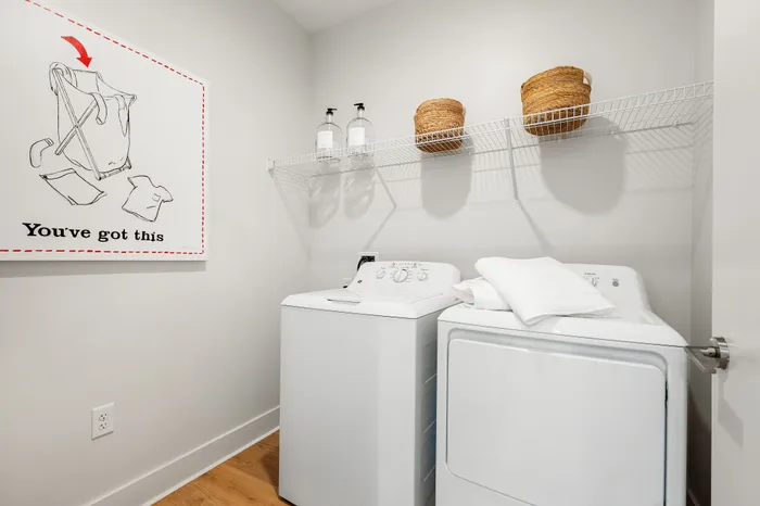 A laundry closet in a The DeSOTA apartment with a washer and dryer, a wire shelf, and a framed art print on the wall.