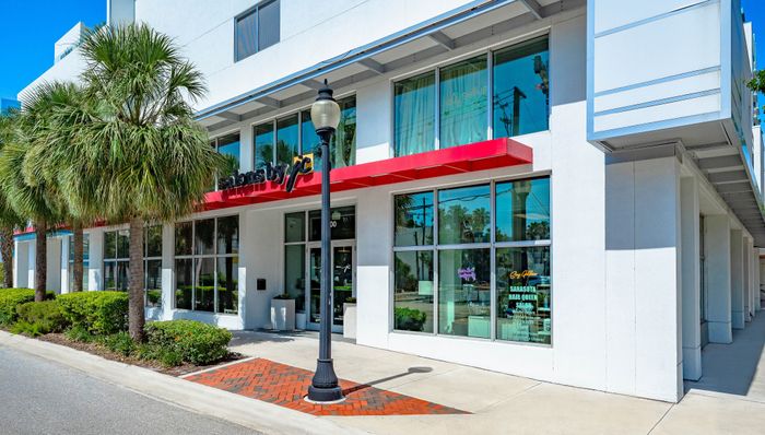 The street-level facade of an apartment building in Sarasota, FL, with a red awning, large windows, and lush palm trees lining the sidewalk.