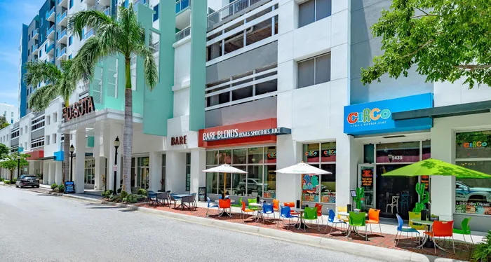The street-level view of The DeSOTA with multiple colorful storefronts, outdoor dining areas, and lush palm trees lining the sidewalk.