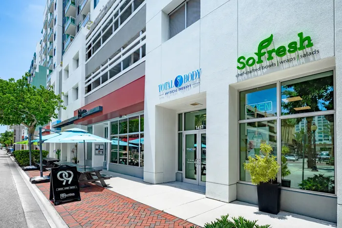 A street-level view of a Sarasota, FL apartment building with multiple storefronts, including a 'So fresh' cafe with outdoor seating and umbrellas.