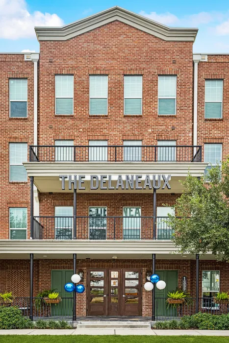 Charming red brick apartment building with elegant balconies, lush greenery, and inviting entryway at The Delaneaux.