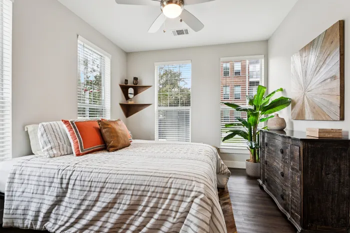 Bright and inviting bedroom with modern decor, featuring a cozy bed, stylish wooden dresser, lush green plant, and large windows for natural light.