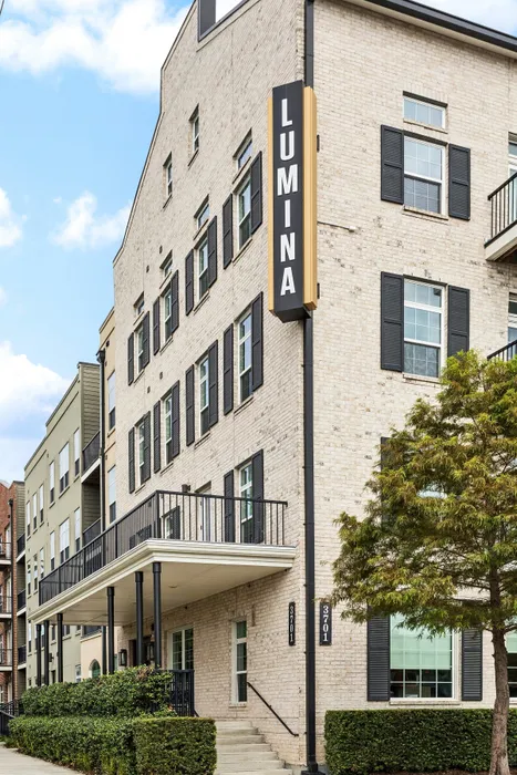 Charming brick apartment complex with modern black accents and a prominent LUMINA sign, set amidst lush greenery.