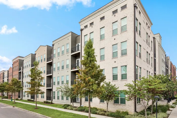 Modern urban apartment complex with stylish brick and siding facade, lush green landscaping, and tree-lined sidewalk.