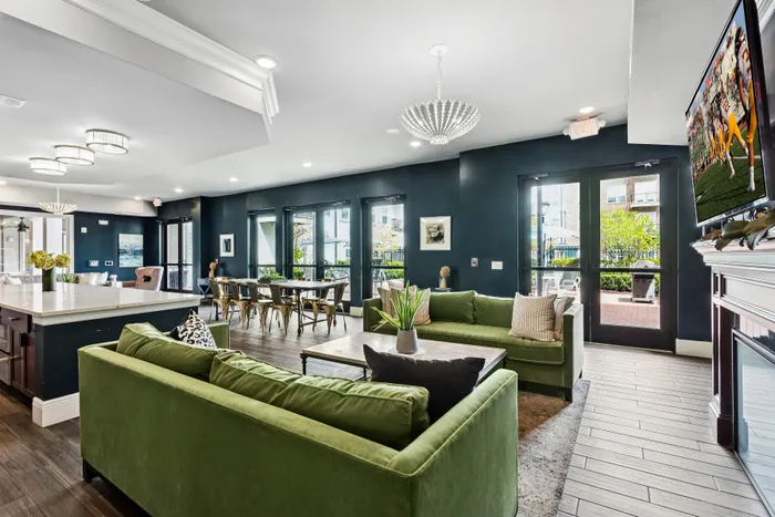 Elegant open-concept living area with stylish green sofas, modern kitchen island, and chic dining space, complemented by abundant natural light and a wall-mounted TV.