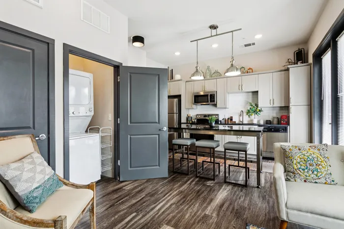 Modern apartment kitchen with sleek white cabinets, stainless steel appliances, and a cozy seating area. Features hardwood floors, a stacked washer and dryer, and stylish lighting.
