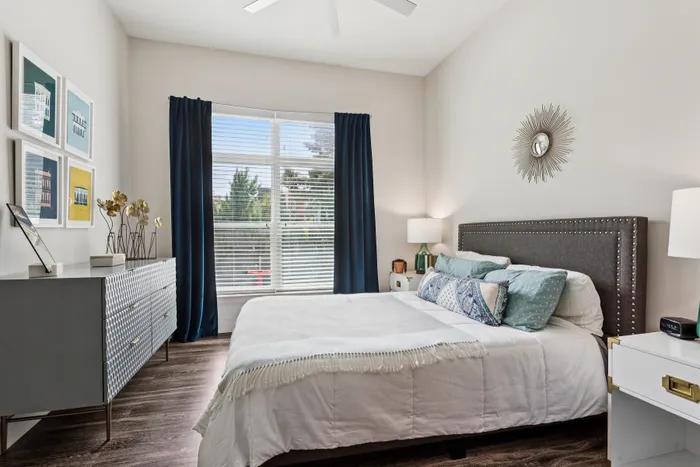 Inviting bedroom with stylish decor, featuring a plush gray headboard, cozy bedding, modern dresser, and large window with navy curtains for natural light.