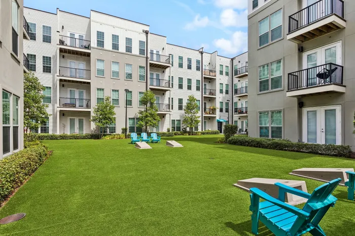 Chic apartment complex featuring a beautifully manicured green courtyard with modern blue seating and corn hole game setups, surrounded by stylish multi-story buildings with balconies.