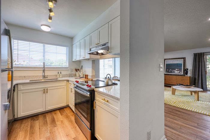 Bright modern kitchen with sleek white cabinetry, stainless steel appliances, and chic wood flooring, leading into a cozy living area with stylish decor.