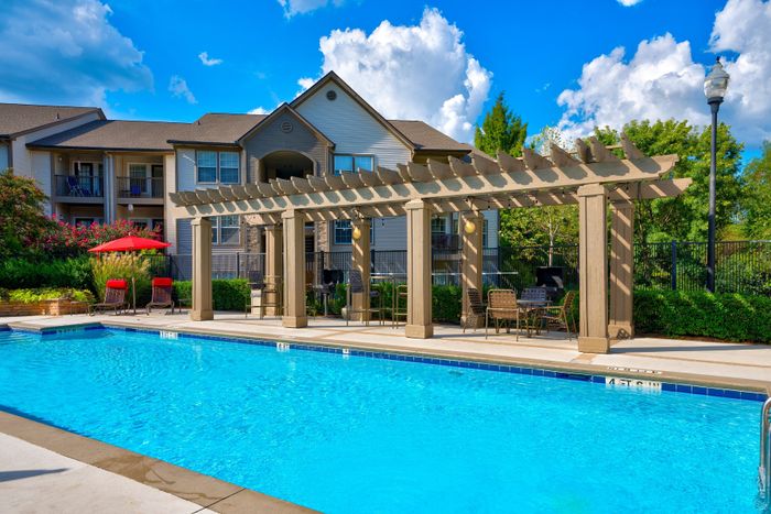 Inviting apartment complex with a sparkling blue pool, stylish pergola seating area, and vibrant landscaping under a clear sky.