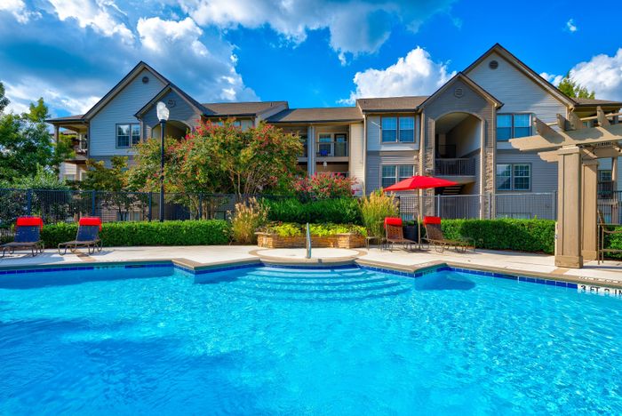 Charming apartment complex with inviting outdoor pool, surrounded by lush greenery and vibrant red umbrellas under a sunny blue sky.