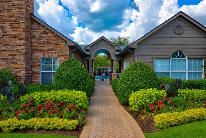 Charming brick and stone apartment entrance with lush landscaping and vibrant greenery leading to a tranquil outdoor pool area.