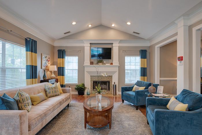 Elegant living room with vaulted ceilings, featuring a stylish beige sofa, blue armchairs, and vibrant yellow and blue curtains. Natural light flows through large windows, highlighting the cozy fireplace and modern decor.