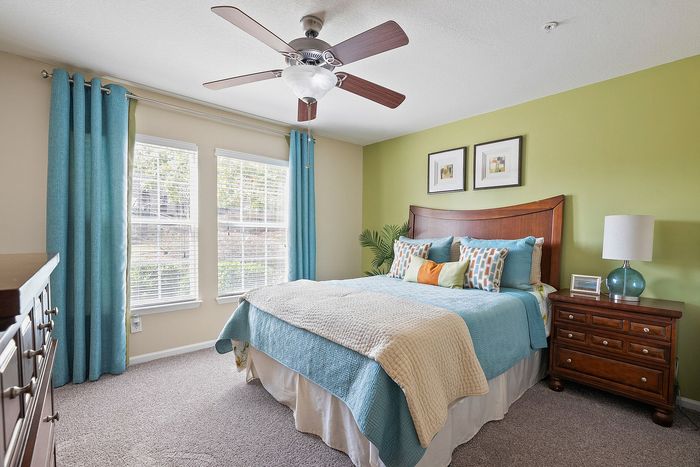 Bright and inviting bedroom featuring a comfortable bed with colorful pillows, elegant wooden furniture, and large sunlit windows with blue curtains.