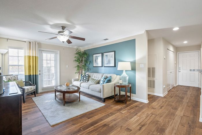 Elegant living room with warm wood flooring, a plush cream sofa accented by vibrant cushions, a modern ceiling fan, and natural light streaming through large windows with stylish curtains.