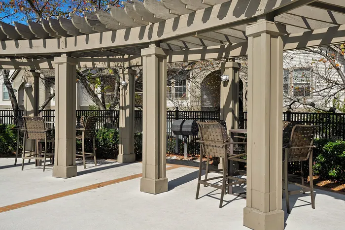 Charming pergola-covered outdoor seating area with elegant high-top chairs and built-in grill, perfect for entertaining or relaxing in a lush garden setting.