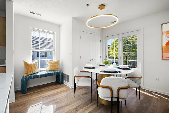 Chic dining area with a modern round table, elegant chairs, and large glass doors opening to a lush view, complemented by a cozy window bench with decorative pillows.
