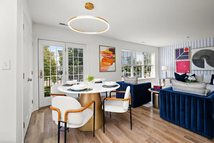 Chic and modern living and dining area with stylish blue sofas, a round dining table, and vibrant artwork, illuminated by natural light through large windows and French doors.