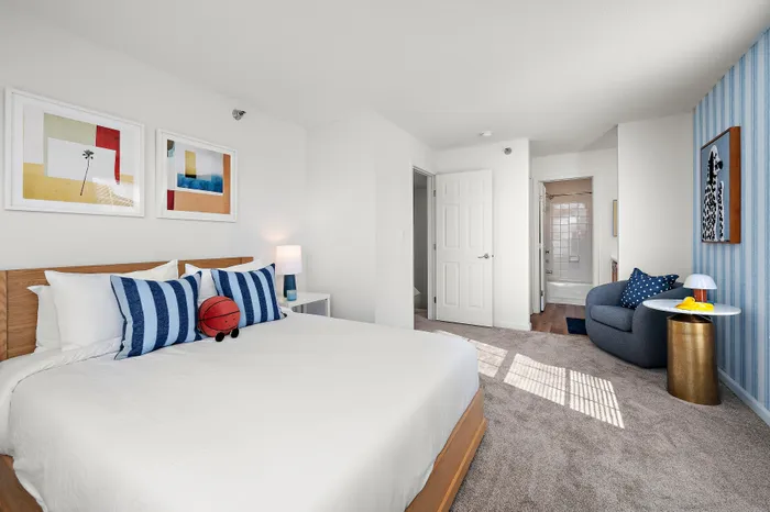 Inviting bedroom with modern decor, featuring a cozy bed with striped pillows, vibrant artwork, and a stylish sitting area. Natural light highlights the elegant neutral palette.