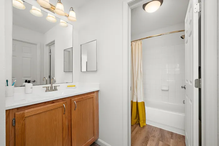 Bright and modern bathroom featuring a wood vanity with ample storage, sleek white countertop, and elegant lighting. Adjacent is a pristine bathtub with a stylish curtain, perfect for a relaxing soak.