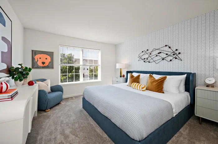 Bright and inviting bedroom with modern decor, plush blue bed, and a large window overlooking greenery. Accented with stylish artwork and cozy furnishing for a serene atmosphere.
