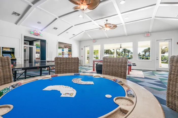 Vibrant recreational room featuring a poker table with cards, a ping pong table, and a view of lush palm trees through large windows.