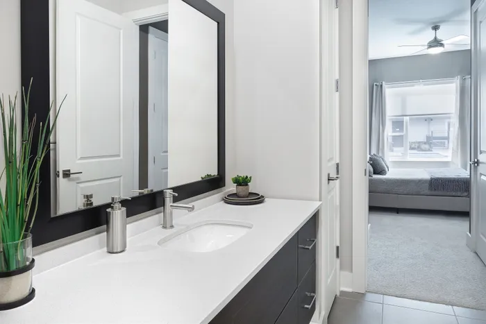 Elegant bathroom with modern fixtures and sleek white countertop, offering a glimpse into a cozy, well-lit bedroom featuring a large window and contemporary decor.