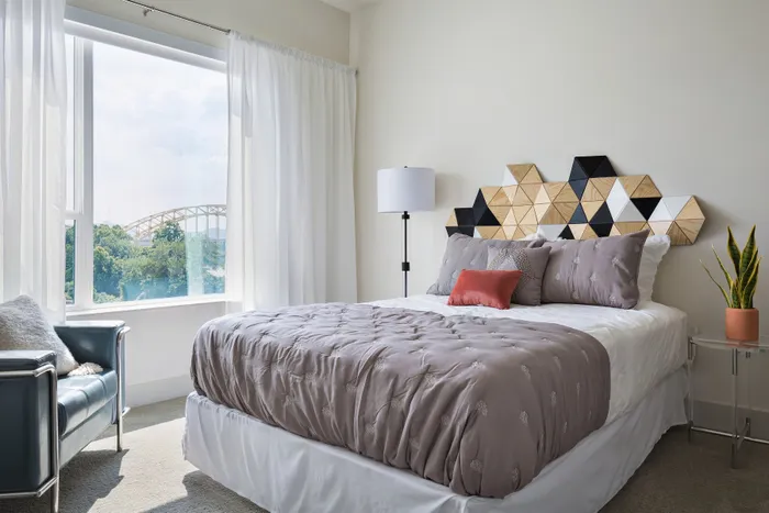 Luxury Apartments For Rent In Strip District, PA - Edge 1909 - A Modern Bedroom With A Unique Wood-And-Black Geometric Headboard, Grey Bedding, And A Large Window Overlooking A Bridge.